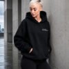 Woman wearing a black oversized Kat Krush hoodie with "Not Loud Just Powerful" chest print, standing against modern concrete architecture with hands in kangaroo pocket.