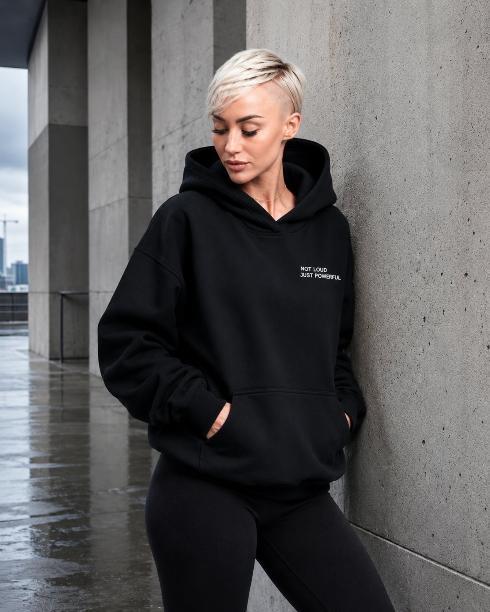 Model wearing a black oversized Kat Krush hoodie and cap beside modern concrete and glass architecture.