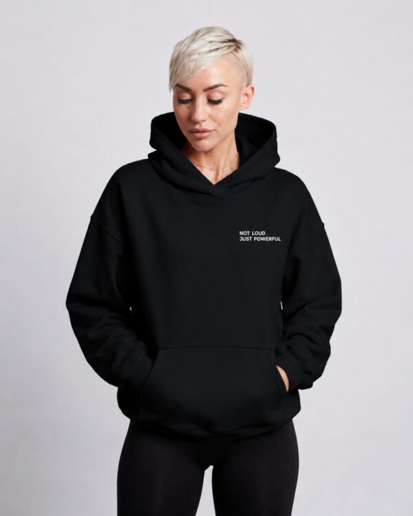 Front studio view of a woman wearing a black oversized Kat Krush hoodie featuring the minimalist "Not Loud Just Powerful" chest print and relaxed heavyweight fit.