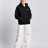 Model wearing a Kat Krush oversized black heavyweight hoodie with "Quiet Confidence." chest print, styled with white cargo trousers in a modern studio setting.