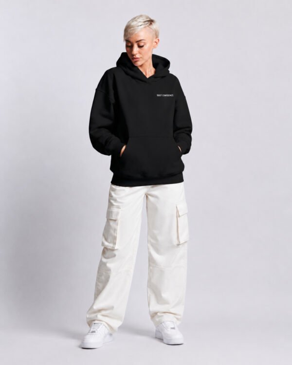 Model wearing a Kat Krush oversized black heavyweight hoodie with "Quiet Confidence." chest print, styled with white cargo trousers in a modern studio setting.