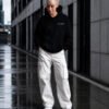 Model wearing a Kat Krush oversized black heavyweight hoodie with "Quiet Confidence." chest print, styled with white cargo trousers in a modern urban city setting.