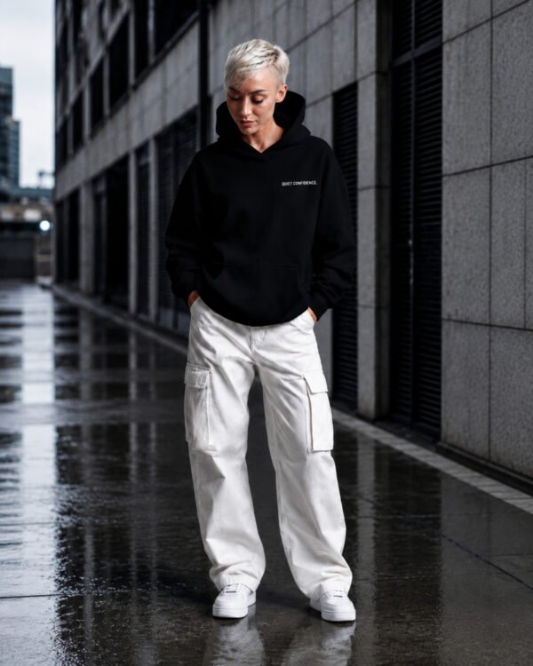 Model wearing a Kat Krush oversized black heavyweight hoodie with "Quiet Confidence." chest print, styled with white cargo trousers in a modern urban city setting.