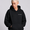 Model wearing a Kat Krush oversized black heavyweight hoodie with "Quiet Confidence." chest print, styled with white cargo trousers in a modern studio setting.