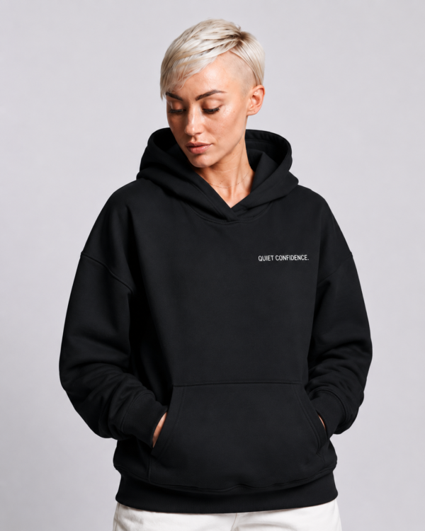 Model wearing a Kat Krush oversized black heavyweight hoodie with "Quiet Confidence." chest print, styled with white cargo trousers in a modern studio setting.