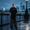 Model wearing a black oversized Kat Krush coordinates T-shirt with DMS back print, standing on the Thames riverside overlooking the London skyline at dusk with St Paul's Cathedral and the City in the background.
