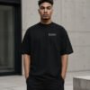 Male model wearing a black Bristol Signature oversized heavyweight T-shirt with white chest coordinates print against contemporary Bristol-inspired architecture.
