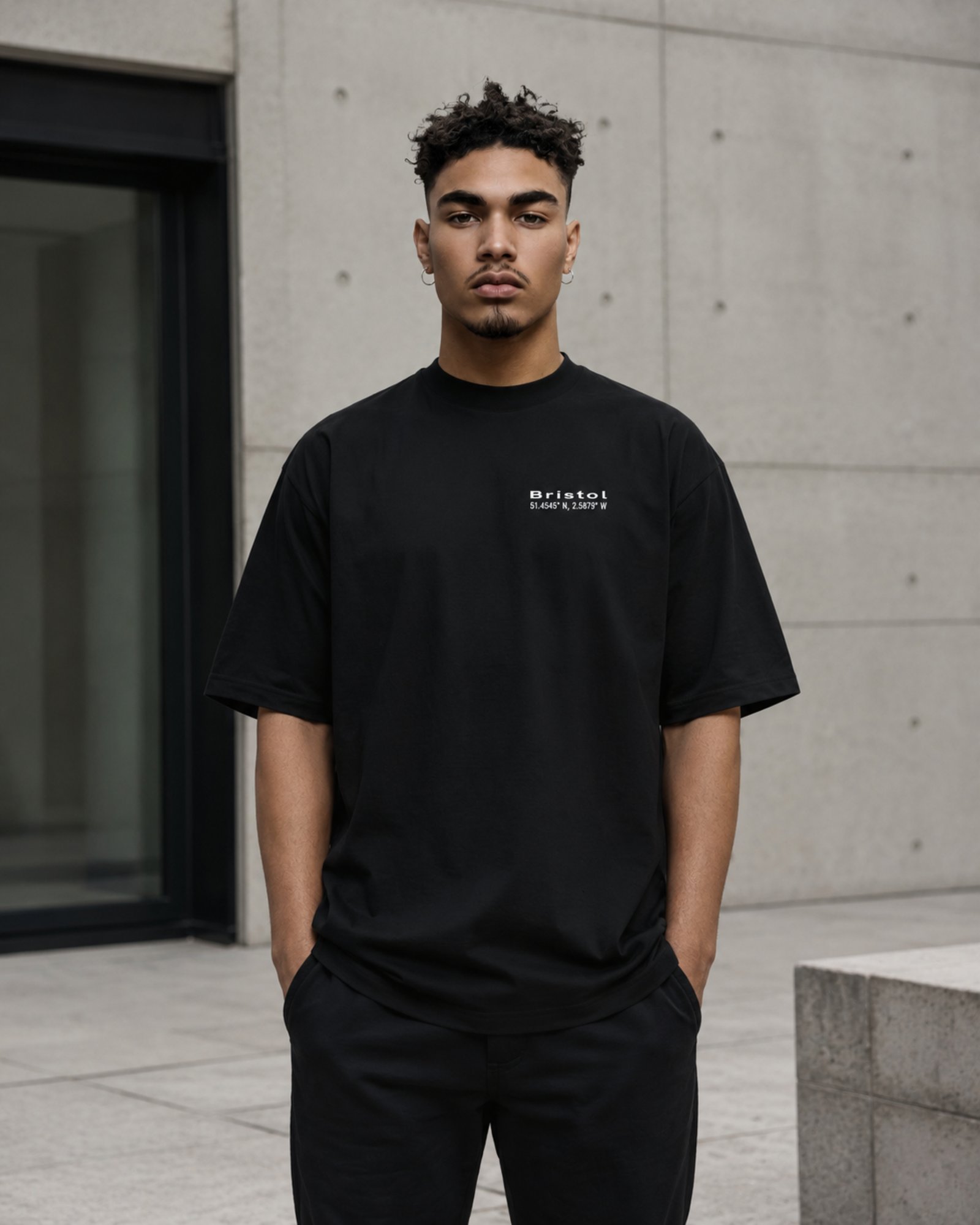 Male model wearing a black Bristol Signature oversized heavyweight T-shirt with white chest coordinates print against contemporary Bristol-inspired architecture.