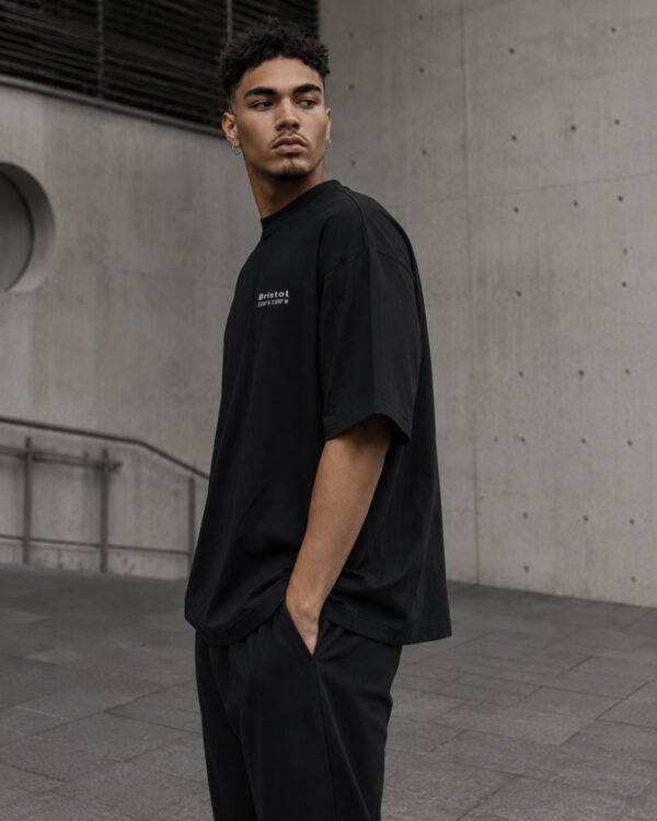 Side profile of a male model wearing a black Bristol Signature oversized heavyweight T-shirt with relaxed fit and subtle white chest coordinates design.