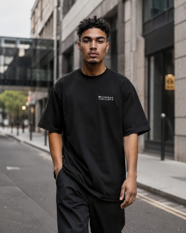 Male model walking through contemporary Bristol-inspired architecture wearing a black Bristol Signature oversized heavyweight T-shirt with minimalist white chest print.