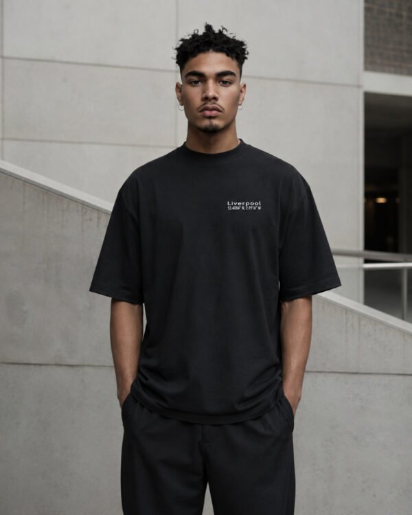 Male model wearing a black Liverpool Signature oversized heavyweight T-shirt with subtle white chest coordinates print against contemporary architecture.