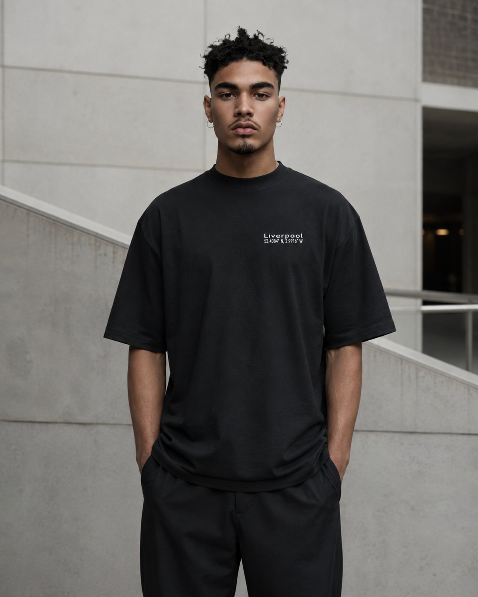 Male model wearing a black Liverpool Signature oversized heavyweight T-shirt with subtle white chest coordinates print against contemporary architecture.