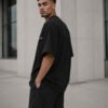 Three-quarter view of a male model wearing a black Liverpool Signature oversized heavyweight T-shirt with subtle white Liverpool coordinates print in a modern architectural setting.