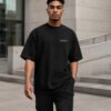 Male model walking through contemporary architecture wearing a black Liverpool Signature oversized heavyweight T-shirt with minimalist white chest coordinates print.