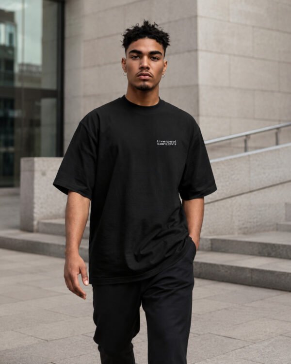 Male model walking through contemporary architecture wearing a black Liverpool Signature oversized heavyweight T-shirt with minimalist white chest coordinates print.