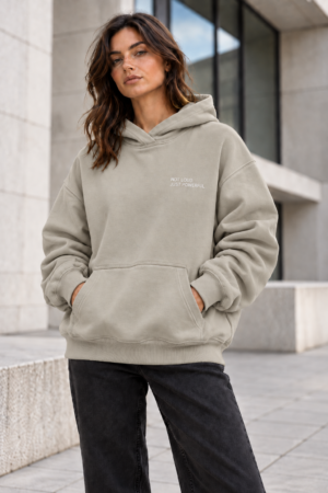 Female model wearing Kat Krush Not Loud Just Powerful stone oversized hoodie with relaxed charcoal trousers against modern concrete architecture.